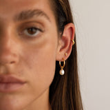 Close-up portrait of a woman wearing gold pearl hoop earrings with a dangling freshwater pearl, paired with a small gold stud and a gold ear cuff on the cartilage. Designed for women who enjoy modern, minimalist jewellery and curated ear stacks, this image shows how pearl hoop earrings can be styled with studs and cuffs for a balanced everyday look or elevated occasion wear. Ideal for those seeking versatile gold pearl earrings that add softness