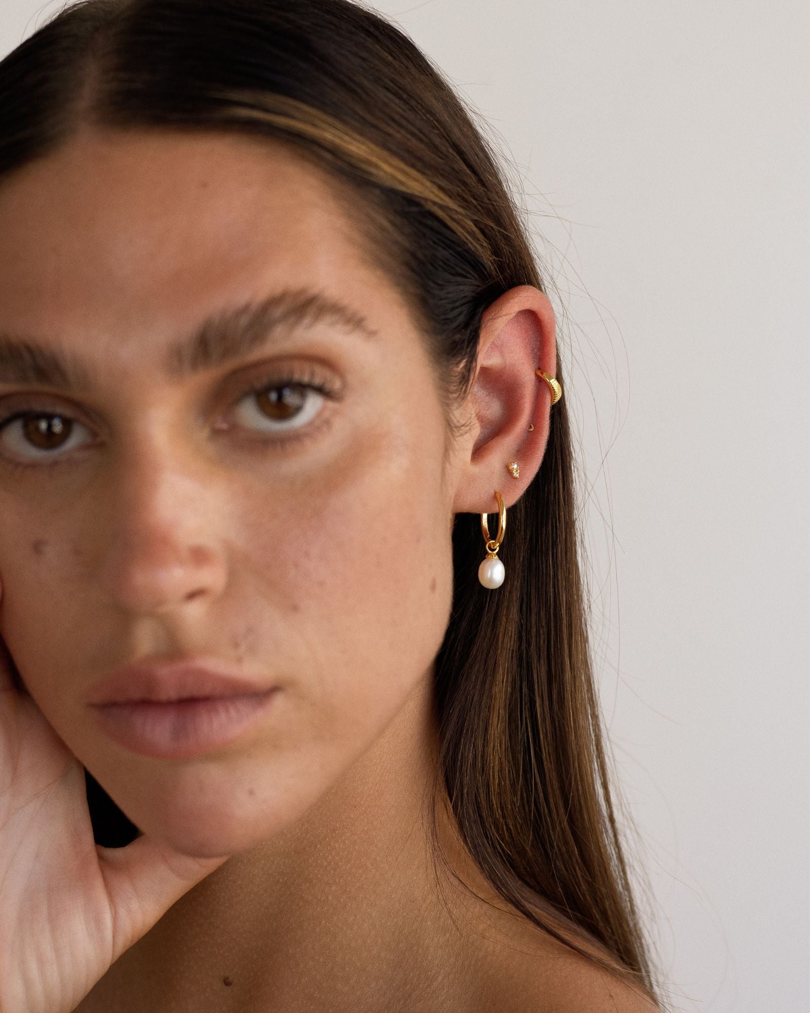 Portrait of a woman wearing small gold hoop earrings with a freshwater pearl drop and a minimalist gold ear cuff, showing an elegant everyday ear stack for women seeking lightweight pearl jewellery, subtle gold accents, and comfortable styling for multiple piercings.