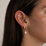 Side profile of an ear styled with a gold flat back stud set with small cubic zirconia stones in the cartilage, a slim gold huggie in the inner ear, and a gold hoop with a pearl drop in the lobe. The image shows a layered ear stack designed for people looking for flat back earrings, cartilage studs, and elegant everyday gold jewellery that offers comfort for sensitive ears and secure wear for long-term styling.