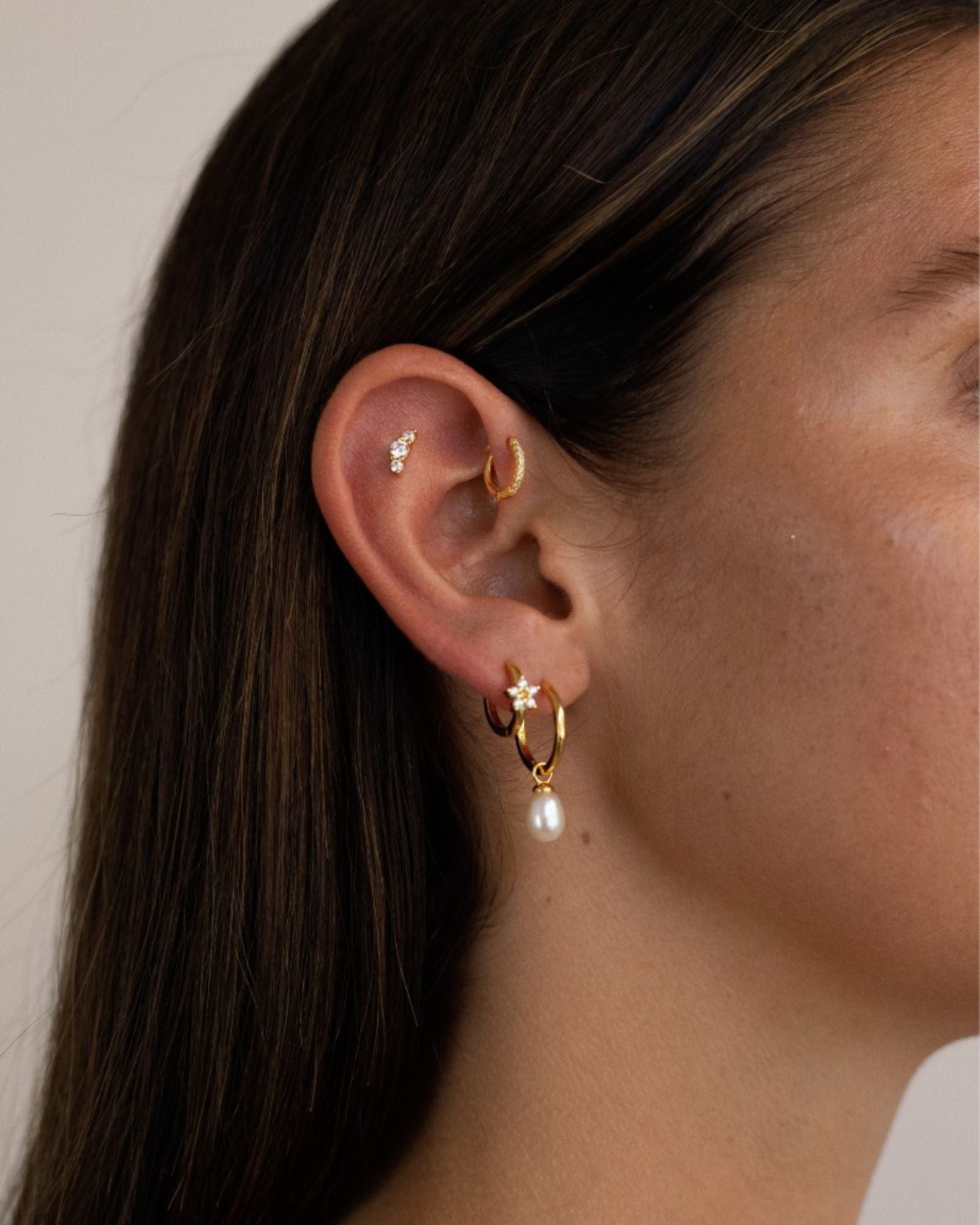 Side profile of an ear styled with a gold flat back stud set with small cubic zirconia stones in the cartilage, a slim gold huggie in the inner ear, and a gold hoop with a pearl drop in the lobe. The image shows a layered ear stack designed for people looking for flat back earrings, cartilage studs, and elegant everyday gold jewellery that offers comfort for sensitive ears and secure wear for long-term styling.