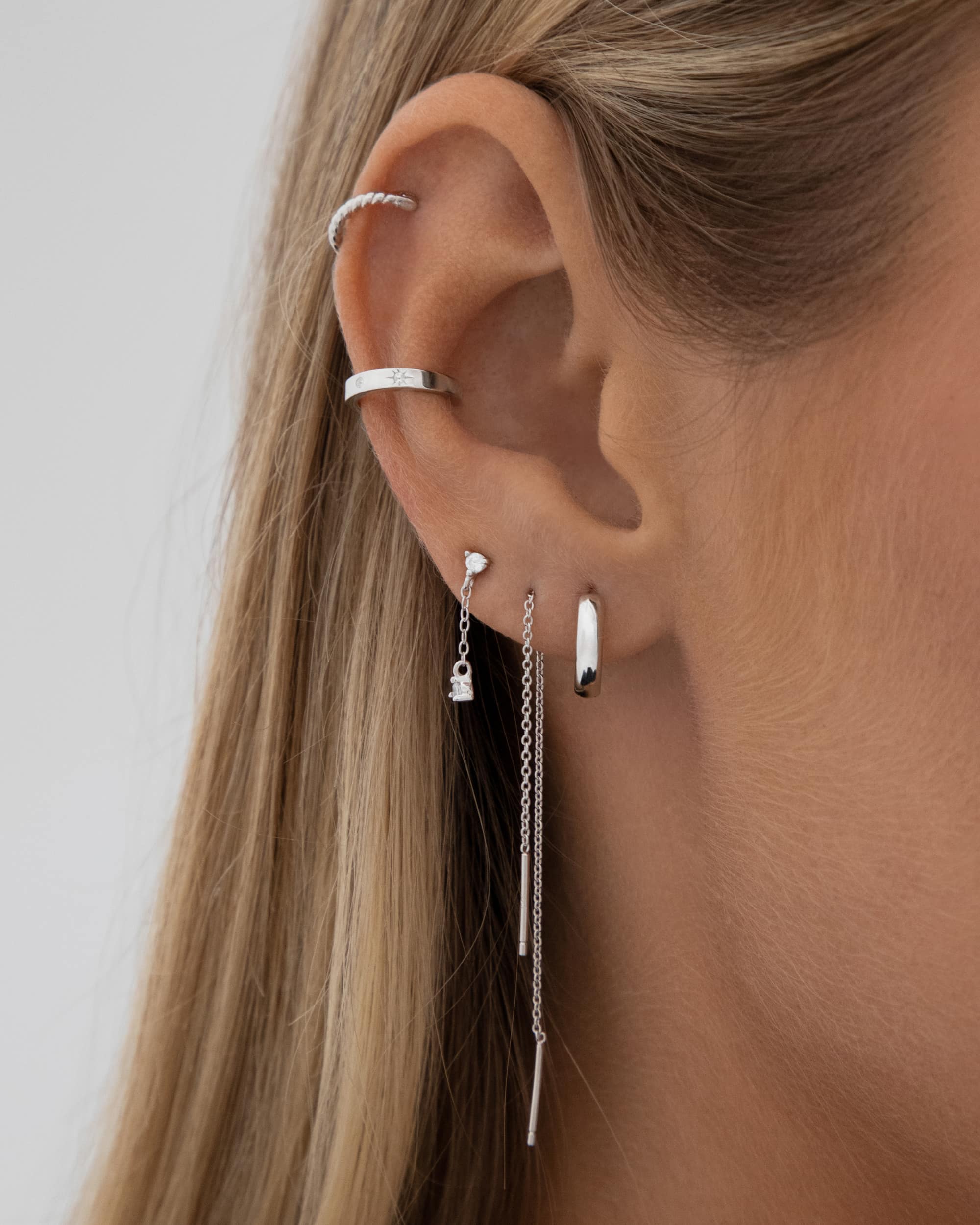 Product image showing a close-up of an ear styled with polished silver jewellery, including a small silver hoop earring, a delicate silver chain drop earring, and minimalist silver ear cuffs along the helix and cartilage. Designed for women seeking modern silver earrings and ear stacking inspiration, this look demonstrates how to layer silver hoops, drop earrings, and ear cuffs for a clean, contemporary aesthetic. The image addresses the need for versatile, hypoallergenic silver jewellery 