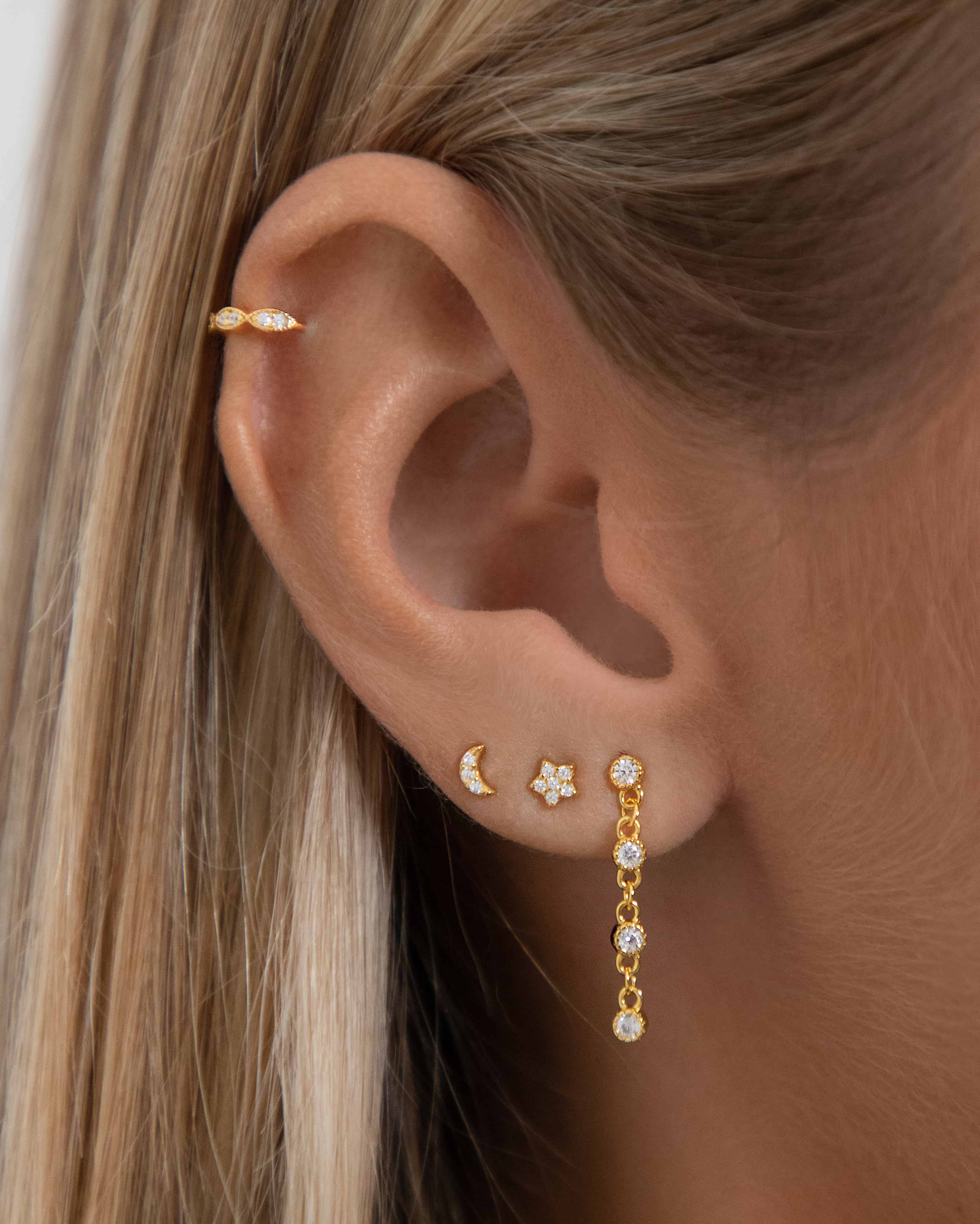 Close-up of an ear wearing gold flat back stud earrings including a crescent moon and star crystal design, paired with a slim gold cartilage hoop and a delicate crystal drop earring in the lobe. Shown on a model to demonstrate how flat back ear piercings sit flush against the ear, ideal for people building a curated ear stack who want secure, low-profile earrings that are comfortable for sensitive ears and suitable for all-day wear and sleeping.