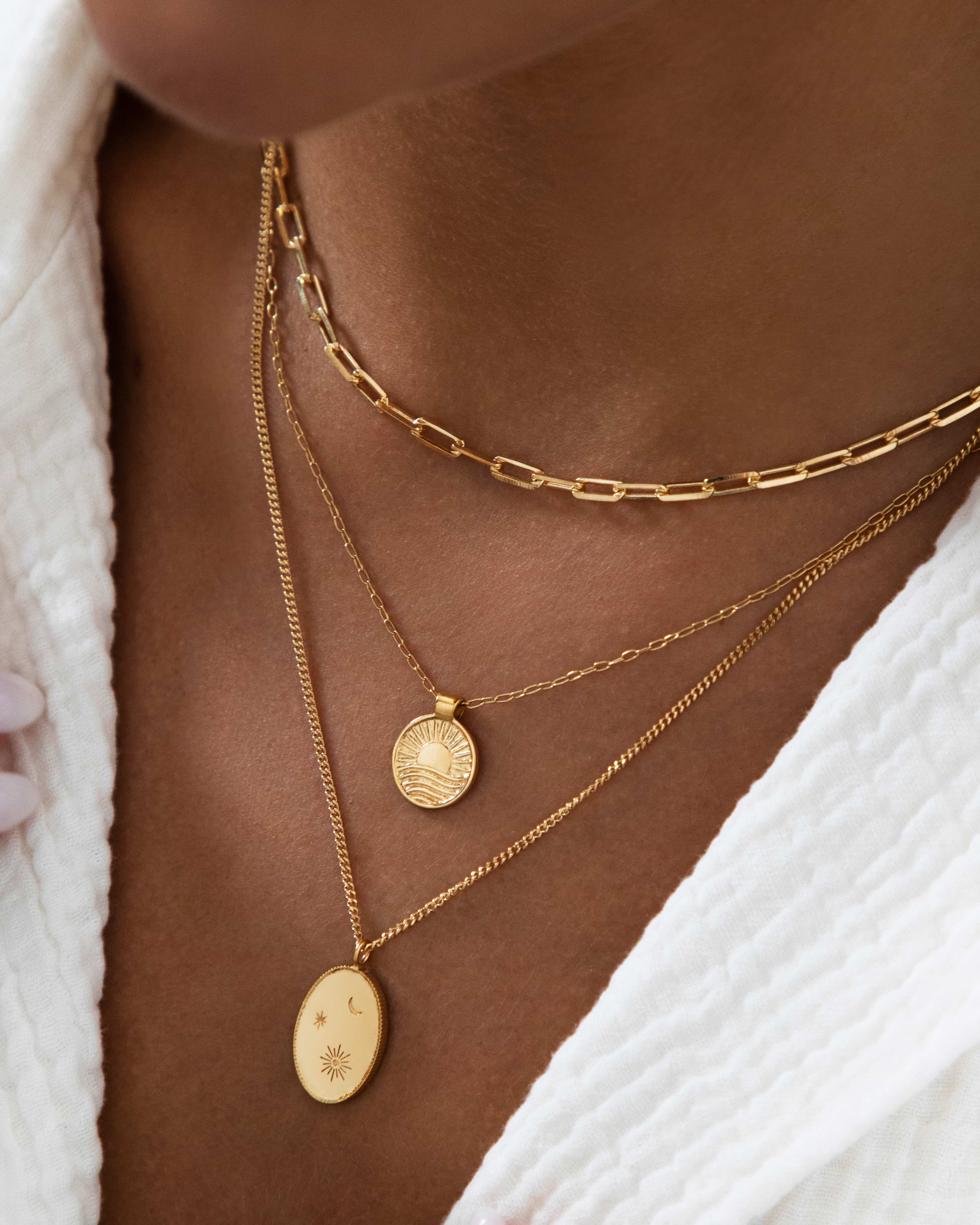 Close-up lifestyle image of a woman wearing a layered gold necklace stack featuring a rectangular link gold chain choker at the collarbone, paired with two fine gold chains with oval sun and celestial coin pendants. Styled on bare skin with a textured white top, the image shows how a gold link necklace anchors an elegant layered look. Designed for women seeking refined gold jewellery and inspiration for effortless necklace layering that feels elevated yet wearable every day.