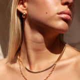 Close-up lifestyle image of a woman wearing a flat gold snake chain necklace layered with a gold paperclip chain and small gold hoop earrings, styled on bare skin in warm natural light. The image shows how a minimalist gold snake chain sits flush on the collarbone when layered with a statement chain. Designed for women seeking refined gold jewellery that layers effortlessly and helps visualise a polished, everyday necklace stack without overcomplicating styling.