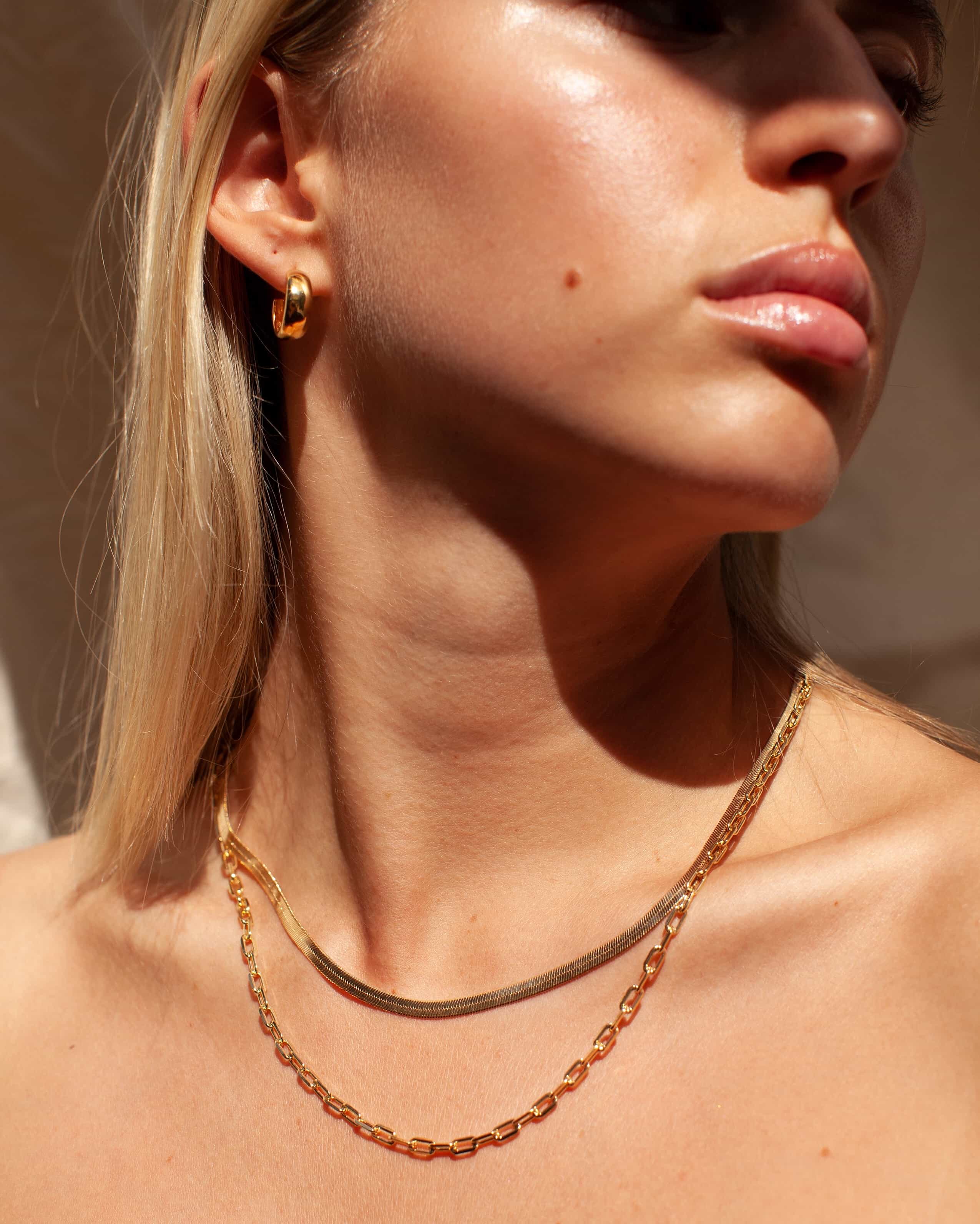 Close-up lifestyle image of a woman wearing a flat gold snake chain necklace layered with a gold paperclip chain and small gold hoop earrings, styled on bare skin in warm natural light. The image shows how a minimalist gold snake chain sits flush on the collarbone when layered with a statement chain. Designed for women seeking refined gold jewellery that layers effortlessly and helps visualise a polished, everyday necklace stack without overcomplicating styling.