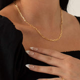 Close-up lifestyle image of a woman wearing a gold rectangular link chain necklace styled alone at the collarbone, paired with a black top and gold ring. The oversized gold link chain creates a clean, structured silhouette that highlights the neckline. Designed for women seeking a modern gold chain necklace that adds effortless polish to everyday outfits and solves the need for a simple yet statement jewellery piece.
