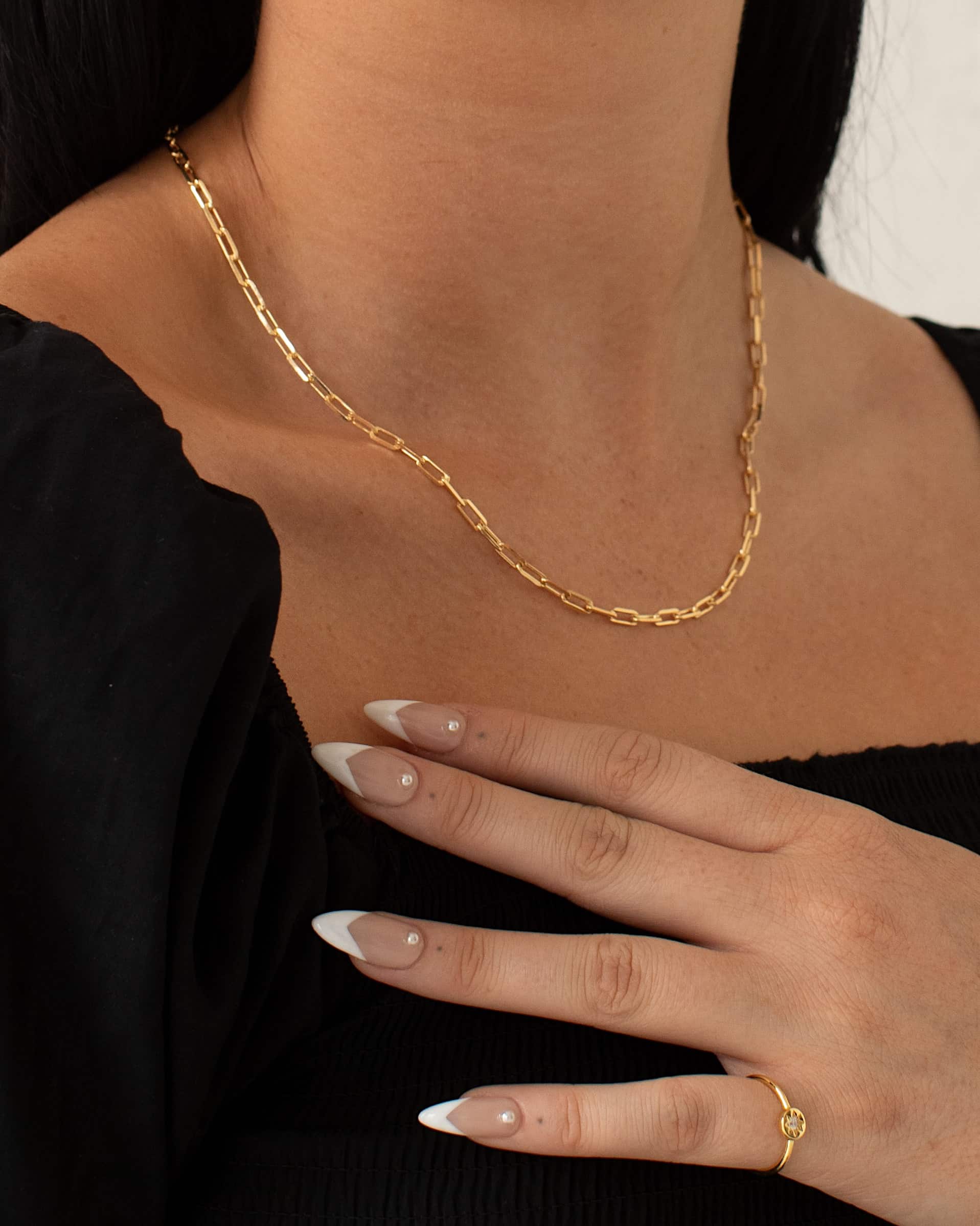 Close-up lifestyle image of a woman wearing a gold rectangular link chain necklace styled alone at the collarbone, paired with a black top and gold ring. The oversized gold link chain creates a clean, structured silhouette that highlights the neckline. Designed for women seeking a modern gold chain necklace that adds effortless polish to everyday outfits and solves the need for a simple yet statement jewellery piece.