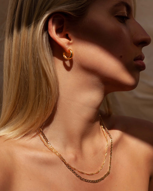Close-up side profile of a woman wearing small polished gold hoop earrings and two layered gold chain necklaces resting along the collarbone. The image highlights minimalist gold jewellery styled on bare skin under warm natural light, emphasizing everyday elegance and layering versatility. Designed for women seeking timeless, understated jewellery that elevates daily outfits and solves the need for durable, easy-to-style pieces suitable for both casual and refined looks.