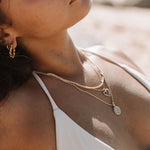 Layered gold necklaces worn on the chest, featuring a delicate gold rope chain paired with fine gold pendant necklaces including a heart charm and a round coin pendant, styled with a white bikini at the beach. This image is for women looking for water-safe gold necklaces that can be worn swimming, sunbathing, or on holiday without removing jewellery. It helps show how lightweight gold chains layer comfortably, resist water exposure, and elevate swimwear with an effortless, everyday luxury look.