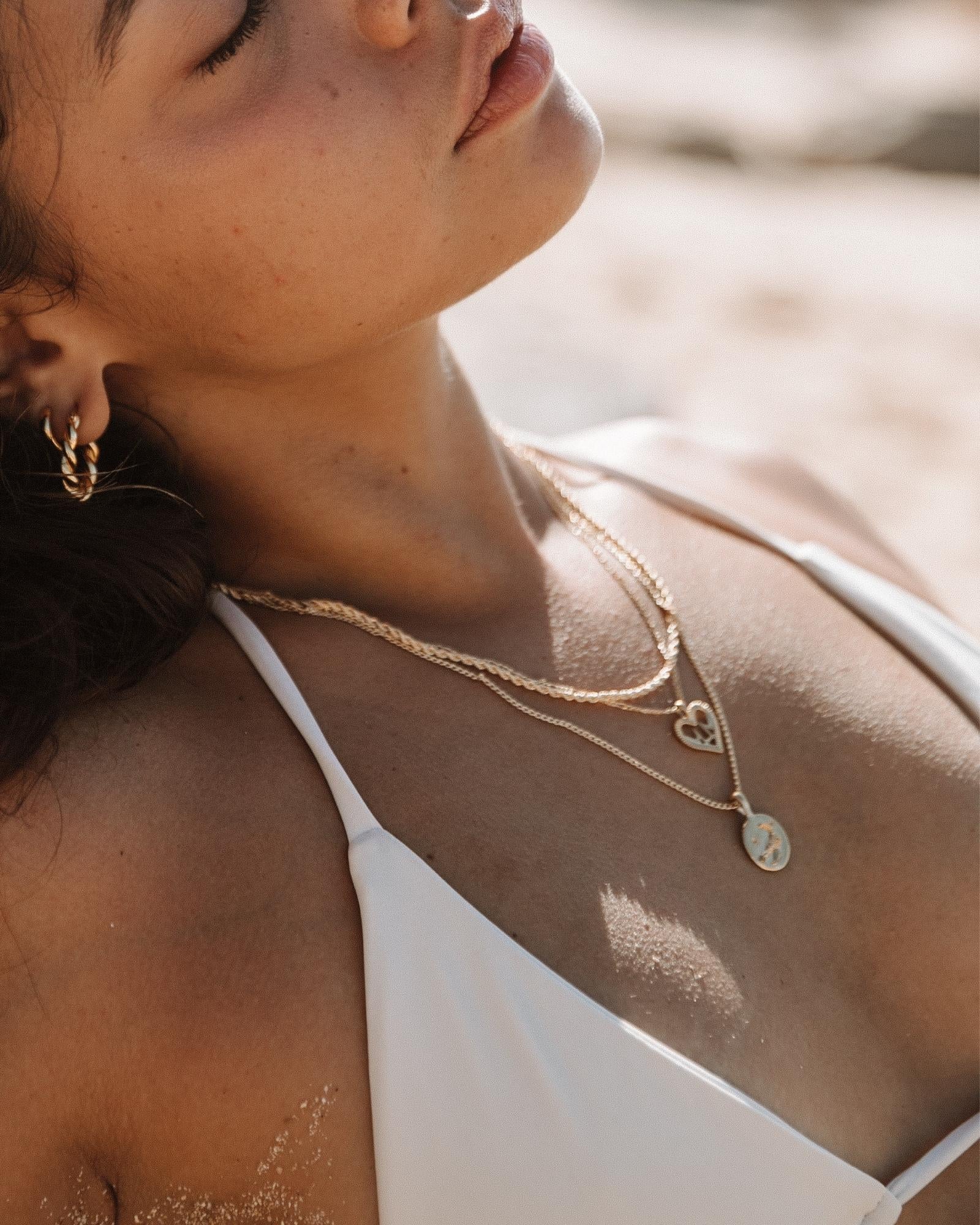 Layered gold necklaces worn on the chest, featuring a delicate gold rope chain paired with fine gold pendant necklaces including a heart charm and a round coin pendant, styled with a white bikini at the beach. This image is for women looking for water-safe gold necklaces that can be worn swimming, sunbathing, or on holiday without removing jewellery. It helps show how lightweight gold chains layer comfortably, resist water exposure, and elevate swimwear with an effortless, everyday luxury look.