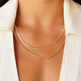 Close-up lifestyle image of a woman wearing a gold snake chain necklace layered with a fine gold rope chain, styled on bare skin beneath an open light shirt. The image shows how a flat snake chain sits flush against the chest when layered, highlighting its smooth, polished finish. Designed for women seeking a minimalist gold snake chain necklace that layers effortlessly and solves the challenge of creating a refined, balanced everyday jewellery stack.