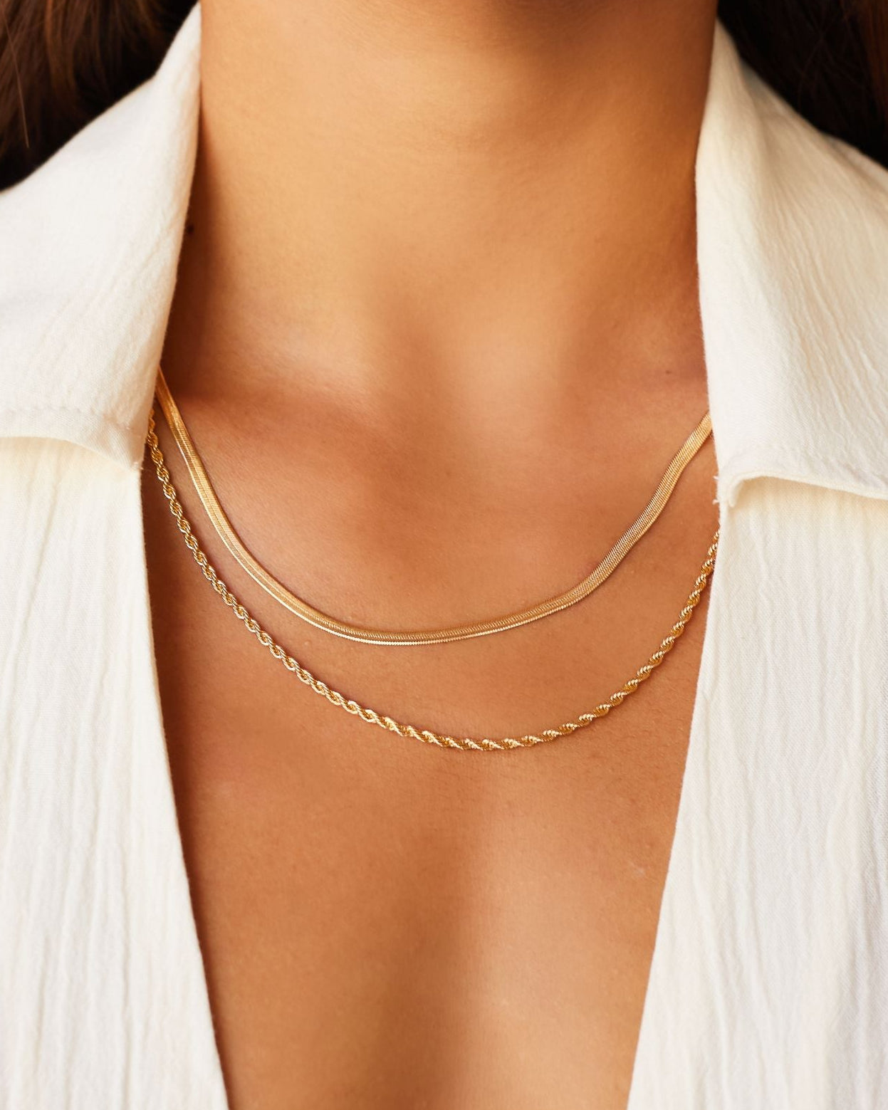 Close-up lifestyle image of a woman wearing a gold snake chain necklace layered with a fine gold rope chain, styled on bare skin beneath an open light shirt. The image shows how a flat snake chain sits flush against the chest when layered, highlighting its smooth, polished finish. Designed for women seeking a minimalist gold snake chain necklace that layers effortlessly and solves the challenge of creating a refined, balanced everyday jewellery stack.