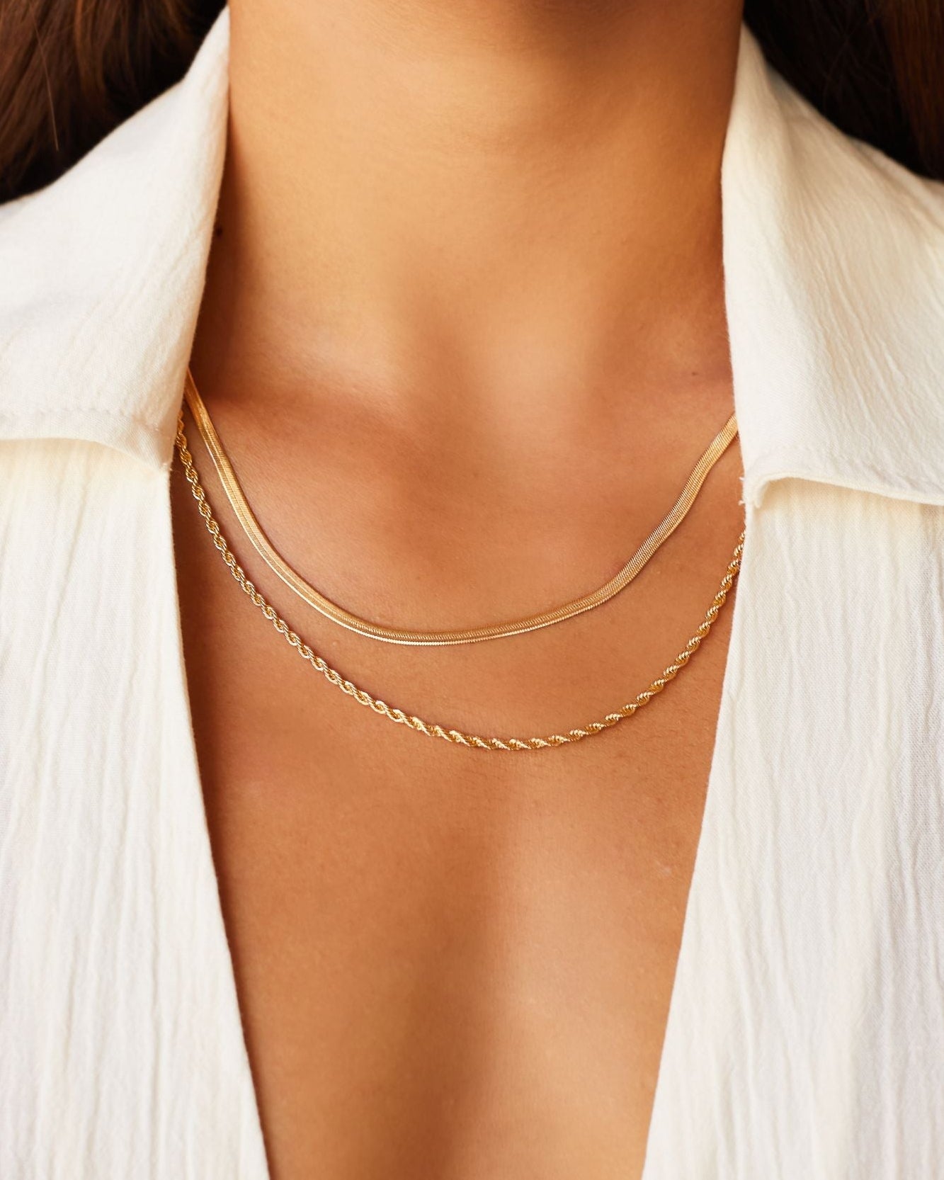 Layered gold necklaces worn on the neck, featuring a slim flat gold chain paired with a delicate gold rope chain, styled on skin with an open white shirt. This image is for women looking for everyday gold necklaces that layer cleanly without tangling and sit comfortably at the collarbone. It helps show necklace length, thickness contrast, and how minimalist gold chains create an elevated, effortless look suitable for daily wear and capsule wardrobes.